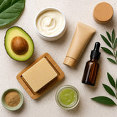 A flat lay of eco-friendly skincare products, including a jar of cream, bar soap on a wooden dish, glass serum bottle, bamboo-packaged tube, avocado half, and green leaves arranged